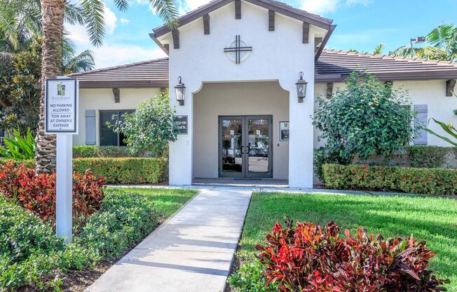 A welcoming building with a light-colored exterior and a prominent cross on the facade. Surrounded by lush greenery, including palm trees and colorful shrubs. Thereâs a clear pathway leading to the entrance, which features large glass doors. Signs indicate parking regulations.
