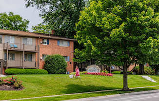 A woman in a pink dress is sitting on a lawn in front of a brick apartment building.