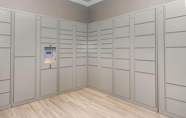 Interior view of a room featuring a wall of gray parcel lockers with a digital keypad and screen. The lockers are neatly arranged in rows, and the floor is made of light-colored wood. The walls are painted in a soft, neutral color, creating a modern and organized space for package delivery.