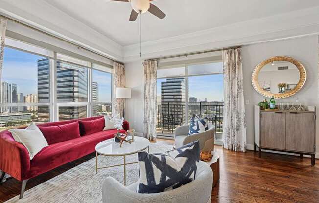 Model living room with a red couch, a white coffee table, and a large window with a view of the city at Dominion Post Oak apartments in Houston, TX