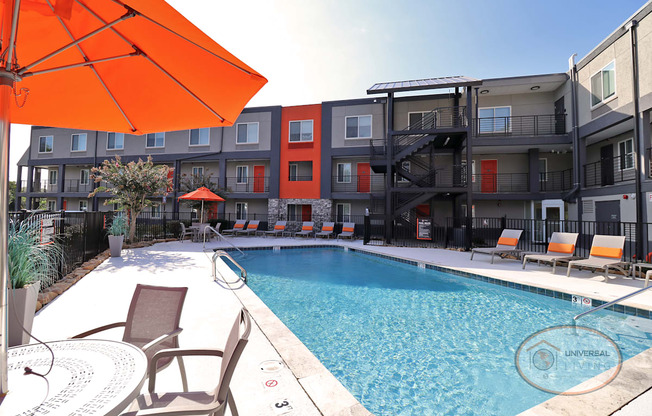 A swimming pool with lounge chairs and tables with orange umbrellas in front of an apartment building.