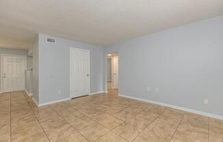 An empty room with light gray walls and tiled flooring. There are two white doors on the left side of the image and an open doorway leading to another room in the background, creating a spacious and clean appearance. The lighting is soft and even, enhancing the neutral tones throughout the space.