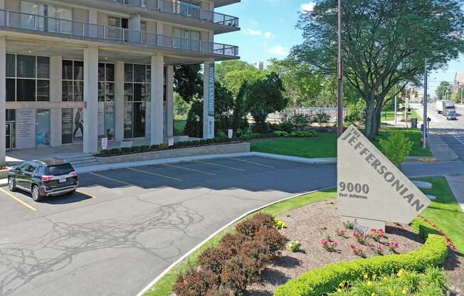 Outdoor image of the Jefferson. The parking lot is empty with the Jefferson sign next to the street. There are green bushes and blue skies.