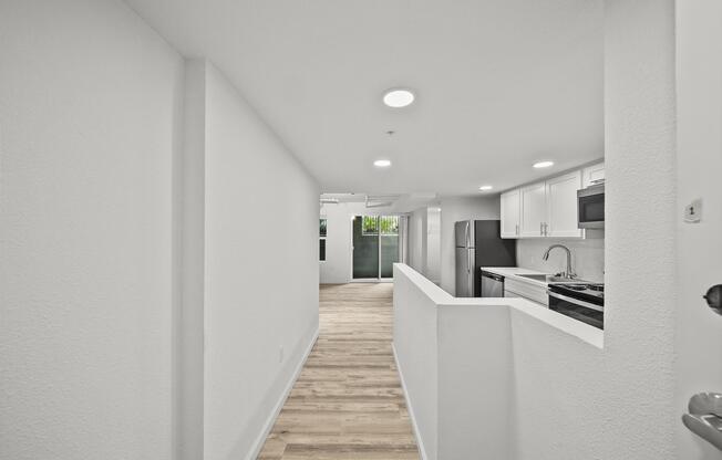 Interior view of a modern, bright hallway leading to an open-plan kitchen and living area. The walls are painted white, with recessed lighting and light wooden flooring. A large window at the end of the hallway allows natural light to fill the space, creating an inviting atmosphere.