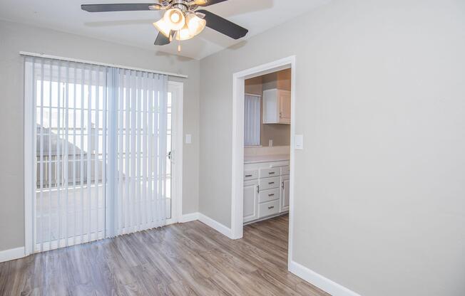An interior view of a room with light gray walls and wood-like flooring. On the left, there are sliding glass doors with vertical blinds leading to an outdoor area. To the right, a doorway leads to a kitchen with white cabinetry. A ceiling fan with five blades is visible in the center of the room.