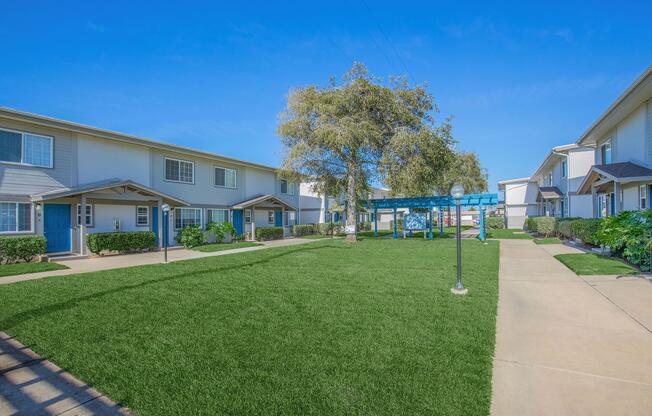 A view of a well-maintained residential complex featuring two-story buildings with blue doors, surrounded by neatly trimmed lawns and shrubs. In the center, a grassy area with a gazebo and a light post under a clear blue sky, creating a serene and inviting atmosphere.