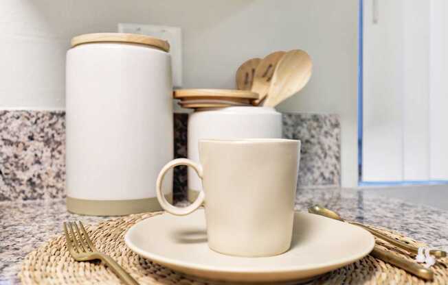 A white mug with a wooden handle is on a plate with a fork and spoon on the table.