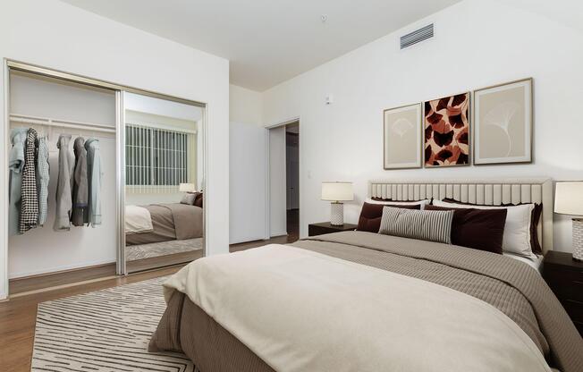 A modern bedroom featuring a large bed with beige and dark brown bedding. There are two bedside lamps on dark nightstands, and a mirrored closet with clothes hanging. The walls are painted white, and two framed artworks are displayed above the bed. A soft area rug covers the wood floor.
