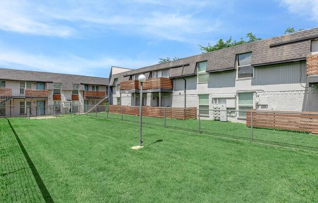A view of a residential apartment complex with multiple two-story buildings. The area features well-maintained grass, fenced yards, and wooden balconies on the upper levels. There's a light post on the lawn, and the sky is clear with a few clouds.