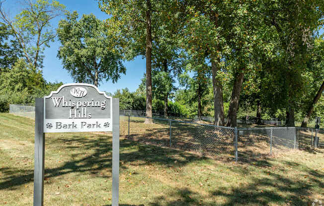 A sign for Whispering Hills Bark Park stands in a grassy area.