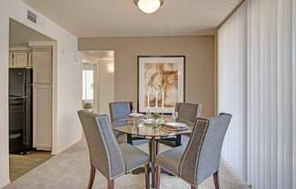 A warm welcoming dining area here at Mirador featuring plush carpet flooring and a round glass-top table surrounded by four upholstered chairs with nailhead trim. A large framed art piece anchors the space, while vertical blinds filter natural light and an open entryway leads toward the kitchen, creating an inviting setting for meals and gatherings.