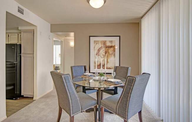A warm welcoming dining area here at Mirador featuring plush carpet flooring and a round glass-top table surrounded by four upholstered chairs with nailhead trim. A large framed art piece anchors the space, while vertical blinds filter natural light and an open entryway leads toward the kitchen, creating an inviting setting for meals and gatherings.