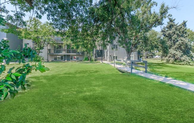 A well-maintained grassy area in front of a multi-story apartment building. The building features balconies, surrounded by trees and greenery, with a clear pathway leading to the entrance. The scene is bright and sunny, suggesting a pleasant outdoor environment.