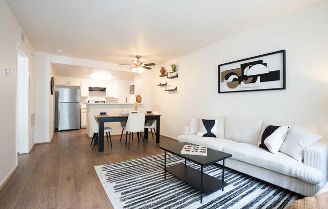 Modern living room and dining area featuring a white sofa, black coffee table, and a striped rug. A dining table with chairs is set beside a well-equipped kitchen with stainless steel appliances. The walls are adorned with minimalist art and decorative shelves. Bright, inviting atmosphere.