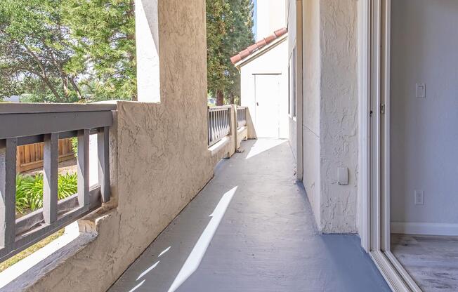 A bright, partially shaded corridor with a smooth concrete floor, flanked by textured walls. To one side, there is a railing overlooking greenery and light filtering through trees. The corridor leads to a white door at the far end, with natural light enhancing the space's openness.