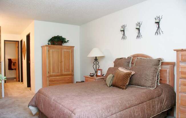 A cozy bedroom here at Eagle Ridge showcasing warm neutral tones with a wood bed frame dressed in textured bedding, matching wood nightstands with a table lamp, soft carpeted flooring, a tall wardrobe for added storage, and wall decor above the headboard, creating a comfortable and inviting retreat connected to a hallway leading toward the bathroom.
