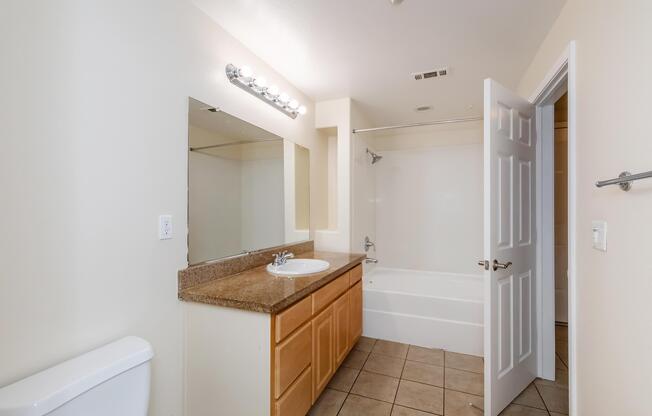 A clean and well-lit bathroom featuring a white bathtub, a beige countertop with a sink, a large mirror, and wooden cabinetry. The walls are painted in neutral colors, and there is a toilet on the left. The space is well-organized and reflects a modern design.