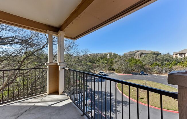 A balcony with a black railing and a concrete floor overlooks a parking lot and a hillside.