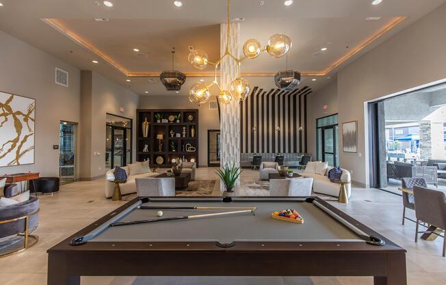 A modern lounge area featuring a billiards table at the forefront, stylish seating arrangements, and contemporary dÃ©cor. The space is well-lit with a large chandelier, and large windows provide views of an outdoor patio area. Decorative shelving and artwork enhance the sophisticated ambiance.