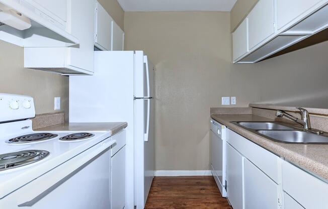 A small kitchen featuring white cabinetry, a white refrigerator, a stovetop with an oven, and a double sink. The countertops are a light color, and the flooring is wood. The walls are a neutral beige, creating a bright, open feel.