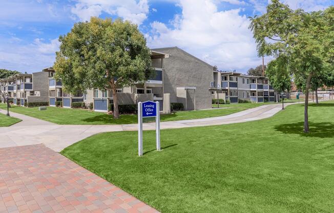 A well-maintained apartment complex with several two-story buildings surrounded by lush green lawns and trees. A sign for the leasing office is prominently displayed in front, along with paved walkways that lead through the landscaped grounds under a partly cloudy blue sky.