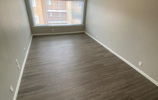 View of room with wood look flooring, ceiling fan, and well lit almost floor to ceiling window.