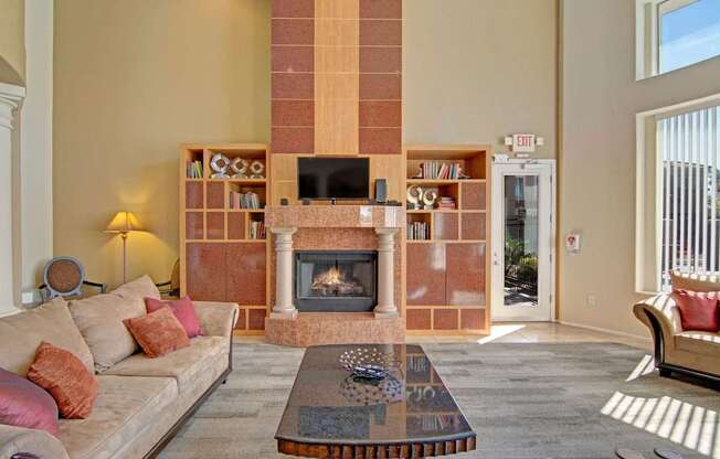 A spacious clubhouse living room here at Dobson Towne Center showcasing a dramatic floor-to-ceiling fireplace with decorative columns and built-in shelving, complemented by a mounted television, plush seating with warm accent pillows, a sleek coffee table, and expansive windows that fill the space with natural light for a comfortable and inviting gathering area.