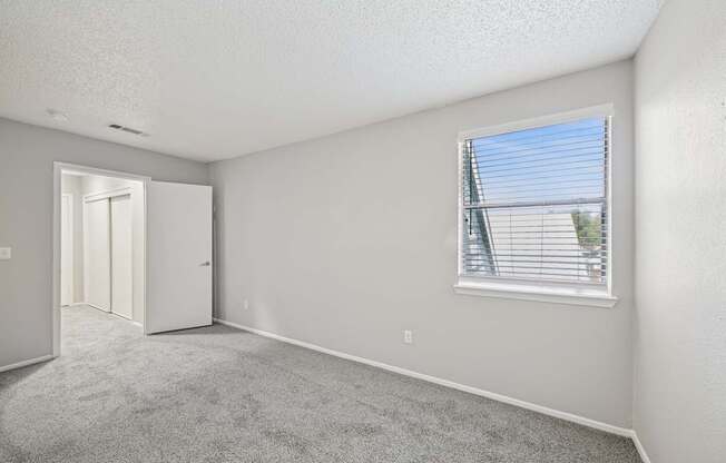 A room with a carpeted floor, a window with blinds, and a door leading to another room.
