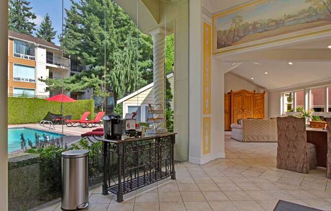 A welcoming community lounge here at Adagio featuring tile flooring, comfortable seating areas, and decorative architectural details, with large windows that overlook a sparkling outdoor pool and red lounge chairs, while a small refreshment station and adjacent gathering spaces create an inviting environment for relaxing, socializing, or enjoying the landscaped courtyard views.