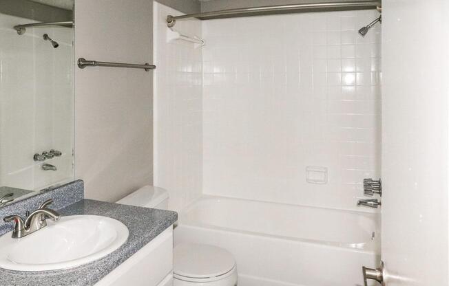A small, functional bathroom featuring a white tiled bathtub with a shower fixture, a large mirror above a sink with a granite countertop, and a toilet. The walls are painted a neutral color and there is a towel bar mounted on the wall. Light reflects off the tiles, creating a bright atmosphere.