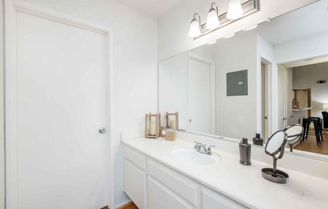 A clean, modern bathroom with a white countertop, double sink, and a large mirror. Soft lighting from three wall-mounted sconces above the sink. A decorative wooden lantern and a small black accessory sit on the counter. A closed door is visible, and a glimpse of a living space can be seen through an open doorway.