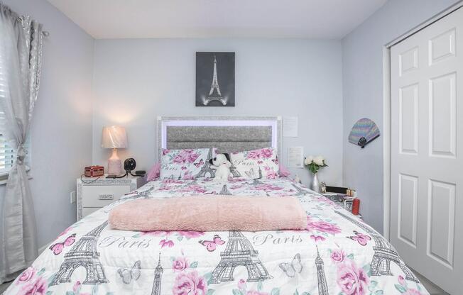 A cozy bedroom featuring a bed with a floral Paris-themed quilt, pink throw blanket, and a stuffed animal. Thereâs a nightstand with a lamp and a flower vase. A wall art of the Eiffel Tower is above the bed, and a closed wardrobe is visible in the background with light-colored walls and curtains.