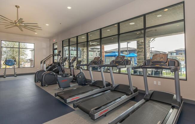 Modern gym interior featuring several treadmills, an elliptical machine, and large windows allowing natural light. The space is well-lit with a ceiling fan, providing a clean and inviting atmosphere for exercising. Outside the windows, there are views of a pool area and landscaped surroundings.