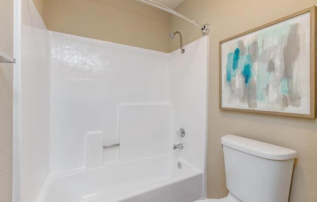 A clean, modern bathroom featuring a white bathtub with a shower area, a toilet, and a painting in a light wooden frame on the wall. The walls are painted in a neutral tone, contributing to a bright and airy atmosphere.