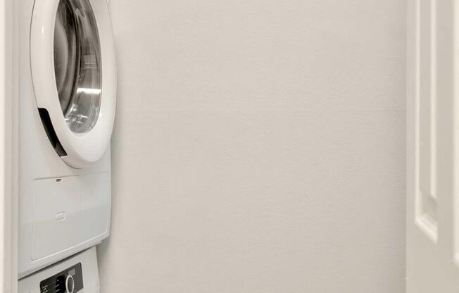 A compact laundry area featuring a white front-loading washer and dryer stacked on top of each other, set against a plain light-colored wall. There is a partially open door to the right, giving a glimpse of the enclosed space.