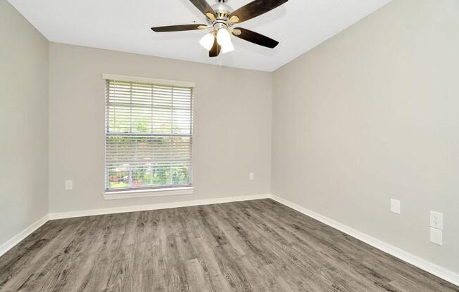 Empty room with light-colored walls and a ceiling fan. A window with blinds allows natural light in, revealing greenery outside. The floor has a wood-like finish, and there are no furnishings in the space, creating a clean and open atmosphere.