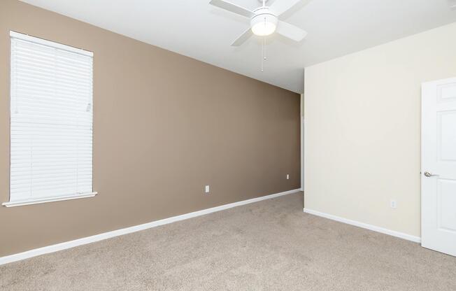 A spacious, empty room featuring beige walls and light-colored carpet. A white ceiling fan is mounted on the ceiling, and there is a window with blinds on one wall. The opposite wall is painted a lighter shade, providing a bright and airy atmosphere.