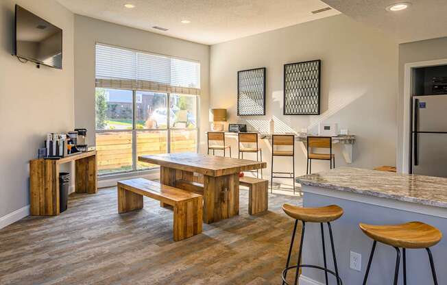 A community gathering room here at Retreat at McAlpine Creek featuring a large wooden dining table with bench seating, bar-height counters with stools, and granite-style countertops. A coffee station and mounted television add convenience, while wide windows with blinds bring in natural light, creating a comfortable space for dining, working, or socializing.