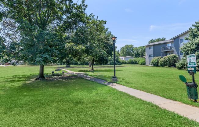 A grassy park area featuring two trees, a concrete path, and a lamppost. There is a picnic table on the grass, and an informational sign is visible nearby. In the background, an apartment building can be seen. The sky is blue with a few clouds.