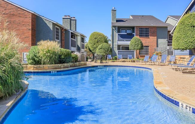 A clear swimming pool surrounded by landscaped greenery and lounge chairs. In the background, residential buildings with a mix of brick and siding are visible under a clear blue sky. The pool area features a shallow end marked with a depth indicator.