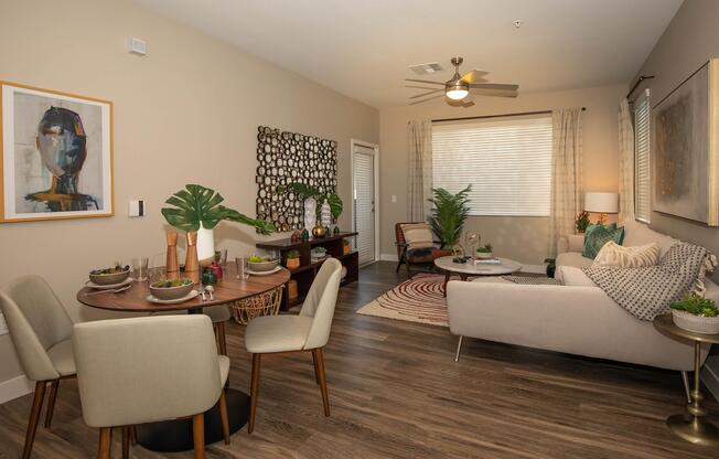 A modern living room and dining area featuring a round dining table set for four, a stylish sofa with cushions, and a coffee table. Decor includes a colorful art piece, a wall decorative display, indoor plants, and a cozy accent chair. Natural light streams through the window, enhancing the inviting atmosphere.