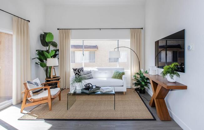 A modern living room featuring a white sofa with decorative cushions, a glass coffee table, and an armchair. The space includes indoor plants, a stylish floor lamp, and a large wall-mounted TV. Natural light floods the room through large windows, creating a bright and inviting atmosphere.