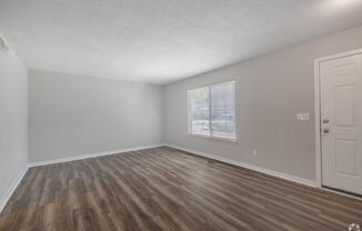 A spacious, empty living room with light gray walls and a large window allowing natural light. The floor features wood-like laminate, and there's a door leading outside. The overall atmosphere is bright and inviting, perfect for personal decoration.