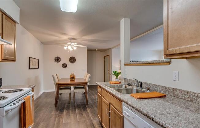 Spokane, WA, Apartments - Canyon Bluffs - Fully Equipped Kitchen with Appliances, Granite Countertops, Wooden Cabinetry, Wood-Style Flooring, Dual Sink, and View of Dining Area with a Table and Chairs.