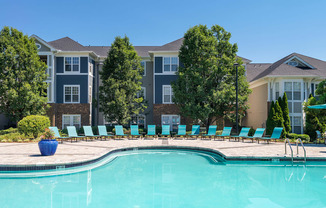 Chapel Hill, NC, Luxury Apartments with In-Unit Washers and Dryers - The Reserve at Meadowmont - Resort-Style Swimming Pool with Sundeck, Lounge Chairs, Surrounding Trees, and Apartment Buildings in the Background.