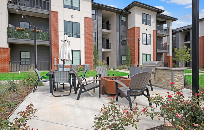 Modern 49 West Apartments courtyard with patio furniture, grill, and fire pit. Surrounding buildings are multi-story with brick and beige exteriors. Lush greenery adds a welcoming feel.