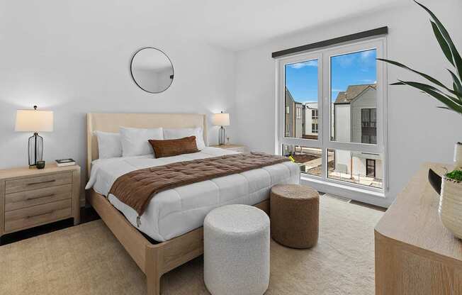 A bedroom with a large bed, a nightstand, and a window overlooking a residential area.