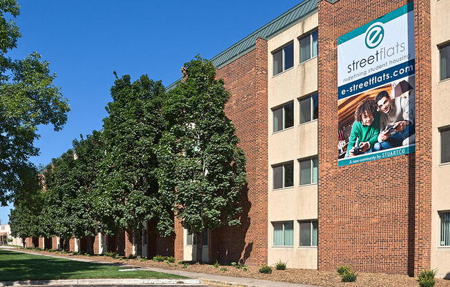 outdoor view of property with tan and brown bricks, tall green trees, and a sign on the building that says "e street flats"