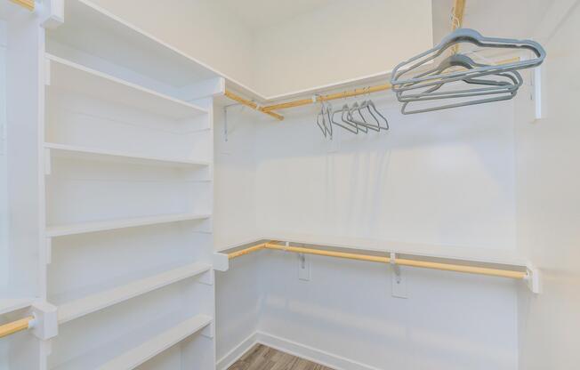 A modern, empty walk-in closet featuring white shelves, a hanging rod with several empty hangers, and a clean, light-colored wall. The floor has a wood-like finish, creating a bright and spacious feel. Ideal for organizing clothing and accessories.
