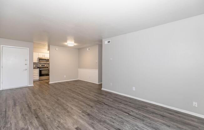 A spacious, empty living area with light grey walls and new laminate flooring. On the left, a white door leads outside. In the background, a modern kitchen is partially visible, featuring stainless steel appliances and white cabinetry. Natural light brightens the room, creating an inviting atmosphere.
